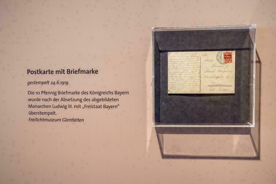 Fotorundgang durch die Sonderausstellung "Eine neue Zeit". Detailansicht des Ausstellungsbereiches "Bayern wird Freistaat". Zu sehen: Postkarte mit Briefmarke, gestempelt 24.6.1919. Die Postkarte wurde nach der Absetzung von Ludwig III. mit "Freistaat Bayern" überstempelt.