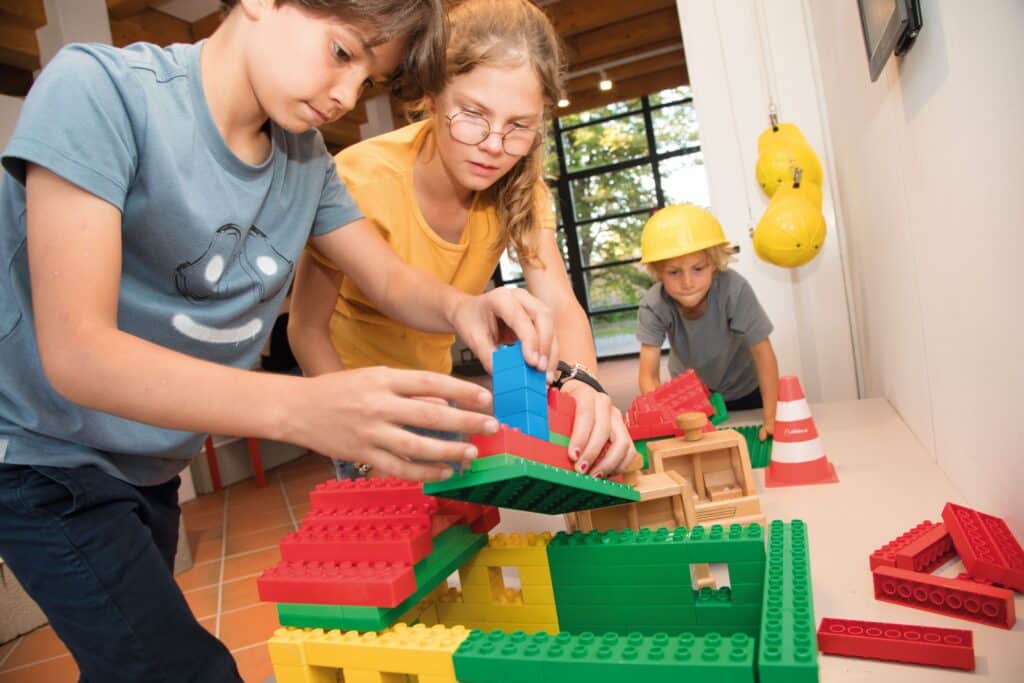Drei Kinder spielen mit Duplo-Steinen. Ein Junge hat einen Bauhelm auf dem Kopf.