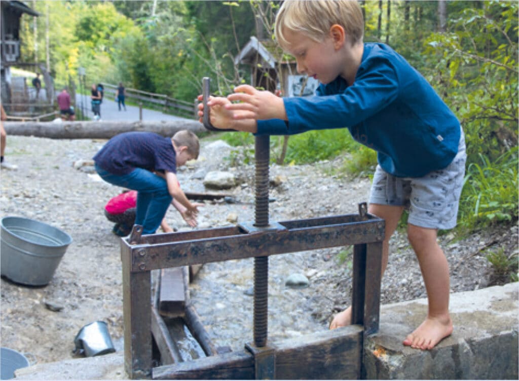 Ein kleines Kind spielt an einem kleinen Wehr, im Hintergrund vergnügen sich weitere Kinder an einem kleinen Bachlauf.