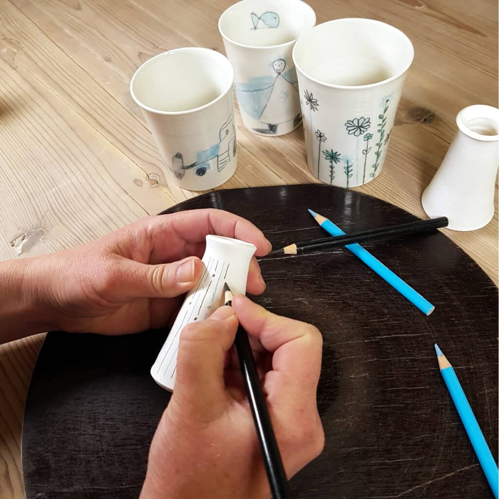 Zeichnen auf Porzellan Mit einem schwarzen Stift zeichnet jemand ein Muster auf eine kleine weiße Vase. Daneben liegen weitere Stifte in Hellblau und Schwraz und dahinter stehen drei bereits bemalte Porzellanbecher.