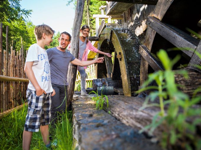 Ein Mann, ein Mädchen und ein Junge stehen an einem wasserbetriebenen Mühlrad aus Holz und betrachten dieses.