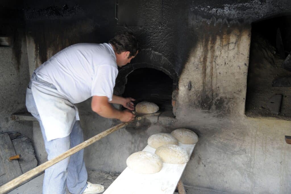 Ein Mann legt einen Brotlaib auf eine Art Schaufel, um ihn in eine Ofenöffnung zu schieben.
