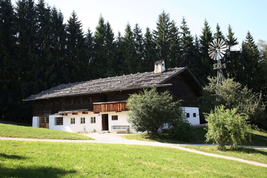 Ein mit Legschindeln gedecktes Haus steht vor einem Nadelwald. Rechts hinter dem Haus befindet sich ein kleines Windrad.
