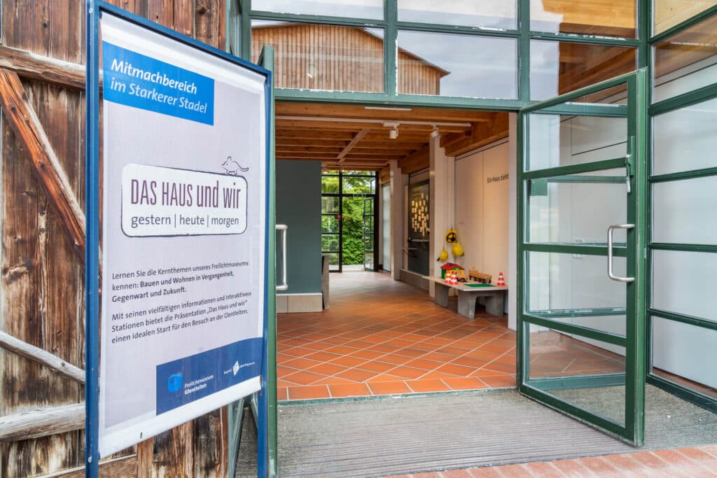 Vor einer geöffneten doppelflügigen Tür steht ein Aufsteller mit einem Hinweis auf "Mimtachbereich im Starkerer Stadel. Das Haus und wir - gestern - heute - morgen".