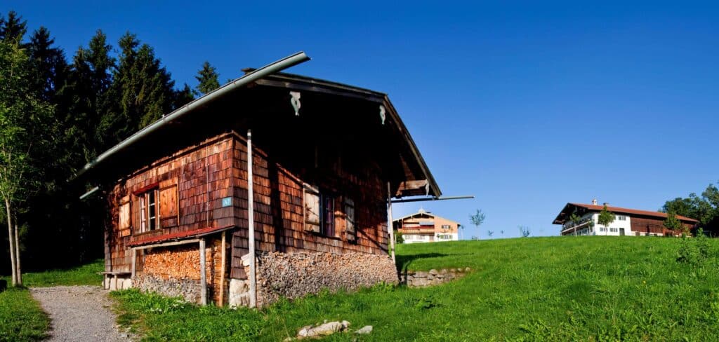 Die Außenwände einer Hütte sind mit Schindeln bedeckt. An den Wänden ist außerdem Brennholz gestapelt. Hinter der Hütte stehen in einiger Entfernung zwei weitere Gebäude.