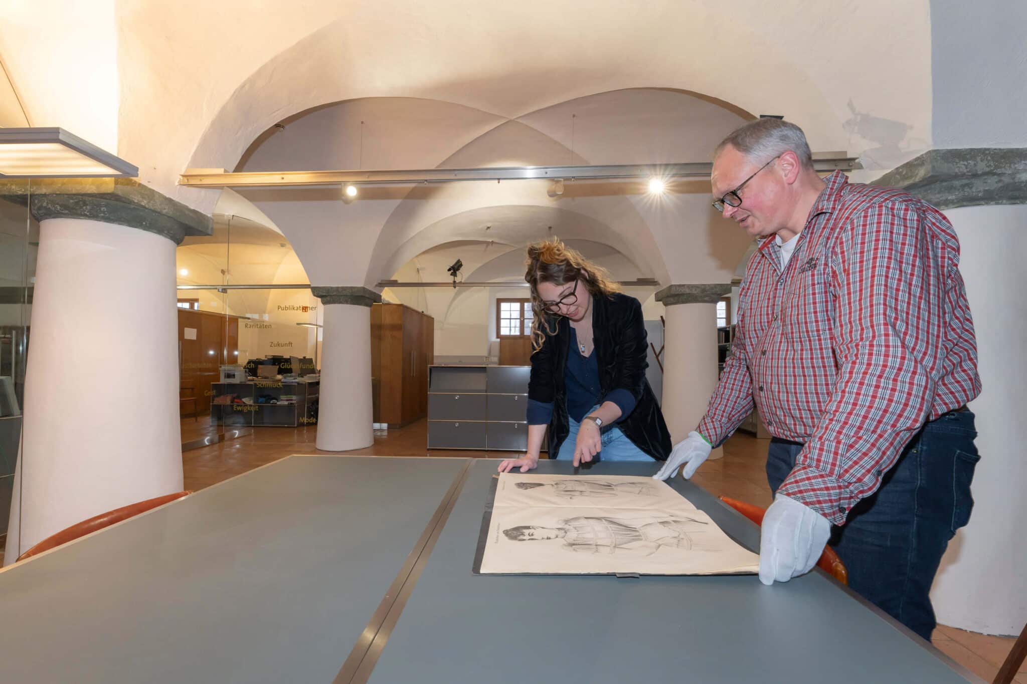 Eine Frau und ein Mann begutachten im Stehen zwei Zeichnungen, die auf einem Tisch liegen. Der Mann trägt dünne weiße Handschuhe.