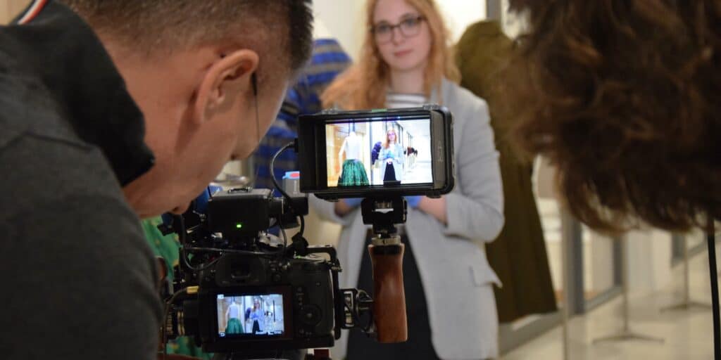 Eine Frau steht vor einer Kamera. Man kann sie in zwei Bildschirmen hinter der Kamera erkennen.