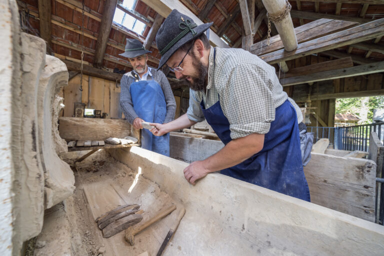 Zwei Männer arbeiten Hand in Hand in einer Werkstatt: Sie schichten vorbehauene kleinere Steine in einen Trog.