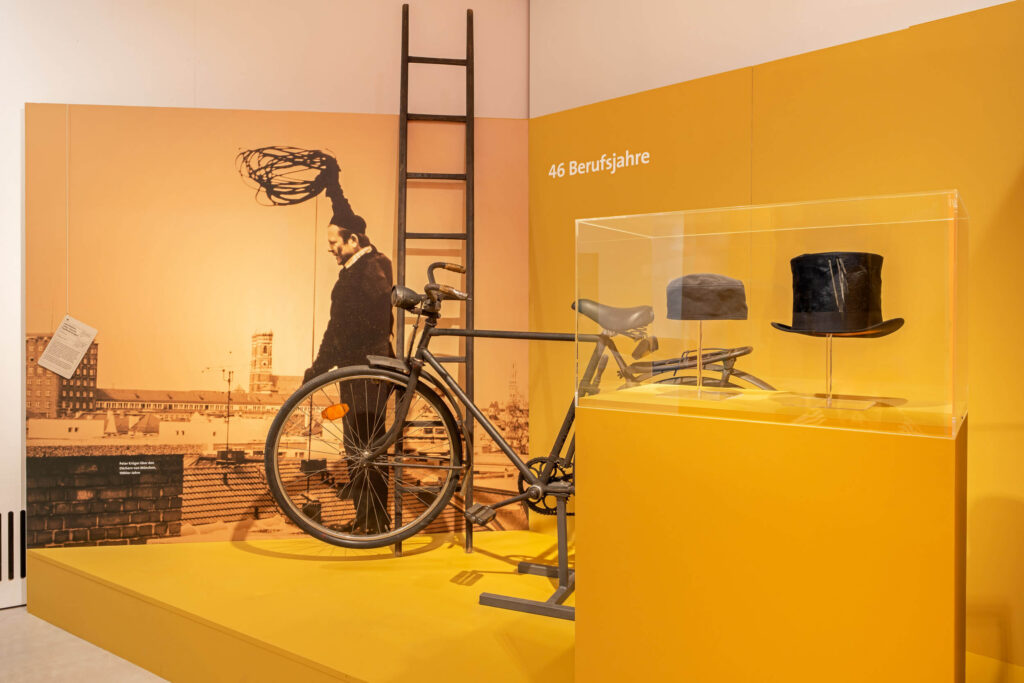 Auf einem ockergelb gestrichenen Podest stehen eine Leiter, ein Fahrrad, eine Kappe und ein Zylinder. Die beiden letztgenannten in einer Vitrine. An der Wand befindet sich ein Foto eines Kaminkehrers auf einem Dach.