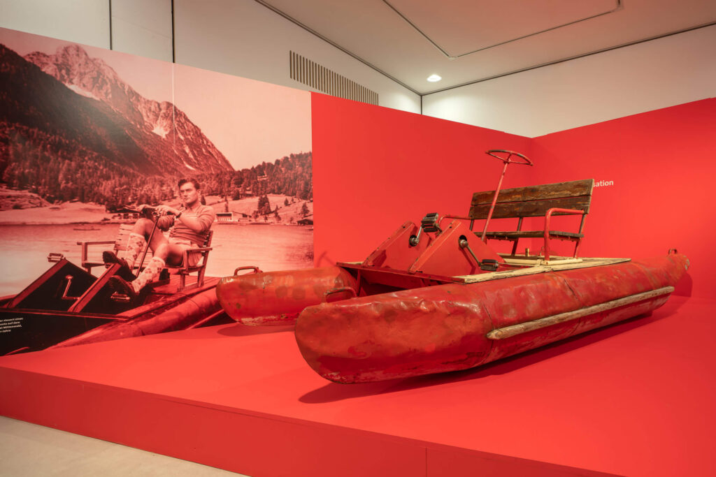 Auf einem rot gestrichenen Podest steht ein ebenfalls rotes historisches Tretboot mit Beulen und Macken. An einer Wand ist ein großes historisches Foto des Tretboots mit Fahrer auf einem See zu sehen.