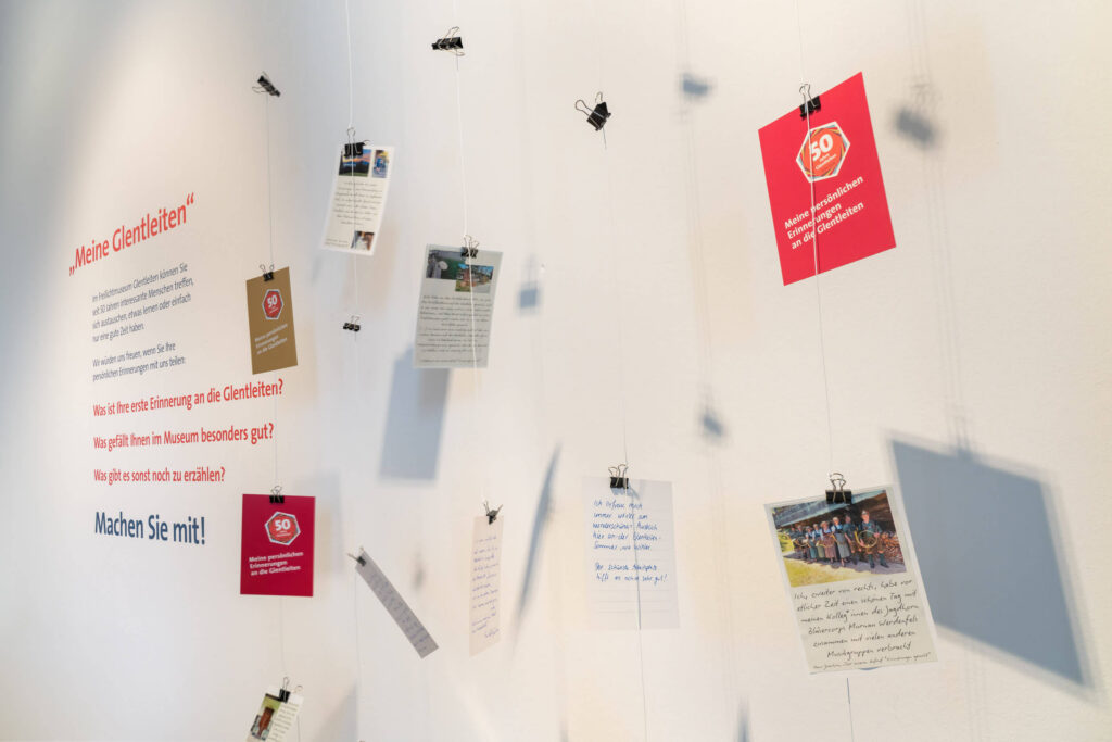 An mehreren Fäden hängen mit kleinen Clips befestigte Postkarten. Links daneben an der Wand steht ein Text, der u.a. heißt: "Meine Glentleiten ... Was ist Ihre erste Erinnerung an die Glentleiten? Was gefällt Ihnen im Museum besonders gut? Was gibt es sonst noch zu erzählen? machen Sie mit!"