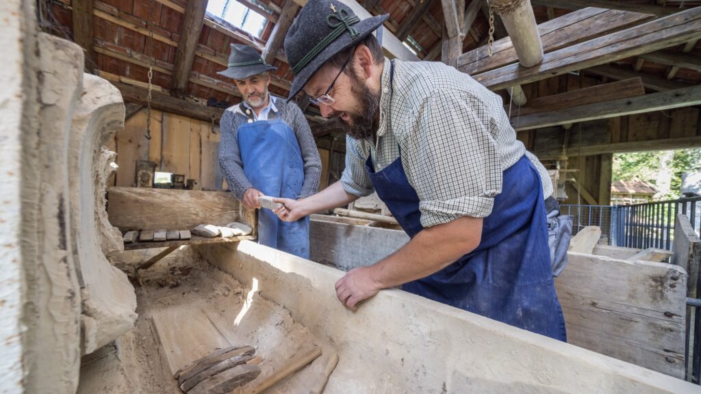 Zwei Männer arbeiten Hand in Hand in einer Werkstatt: Sie schichten vorbehauene kleinere Steine in einen Trog.