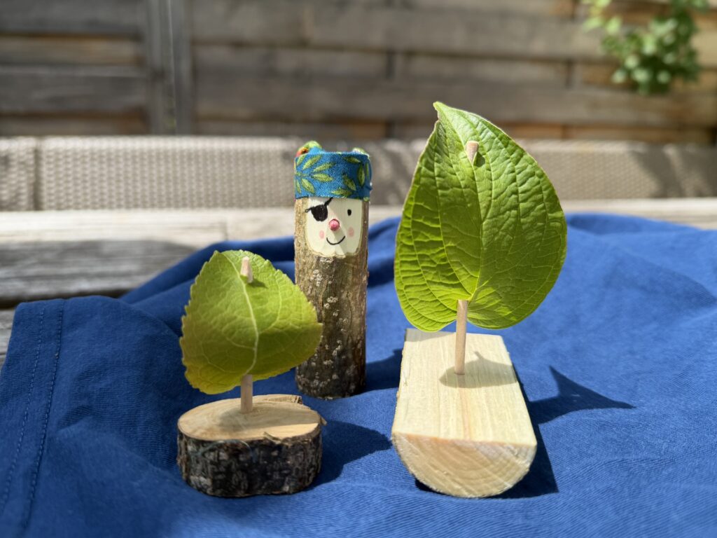 auf einem blauen Stoff stehen zwei aus je einem Holzstück, einem kleinen Pfahl und einem Blatt als Segel gefertigte Bötchen. Ebenfalls aus Holz komplettiert ein Pirat das Bild.