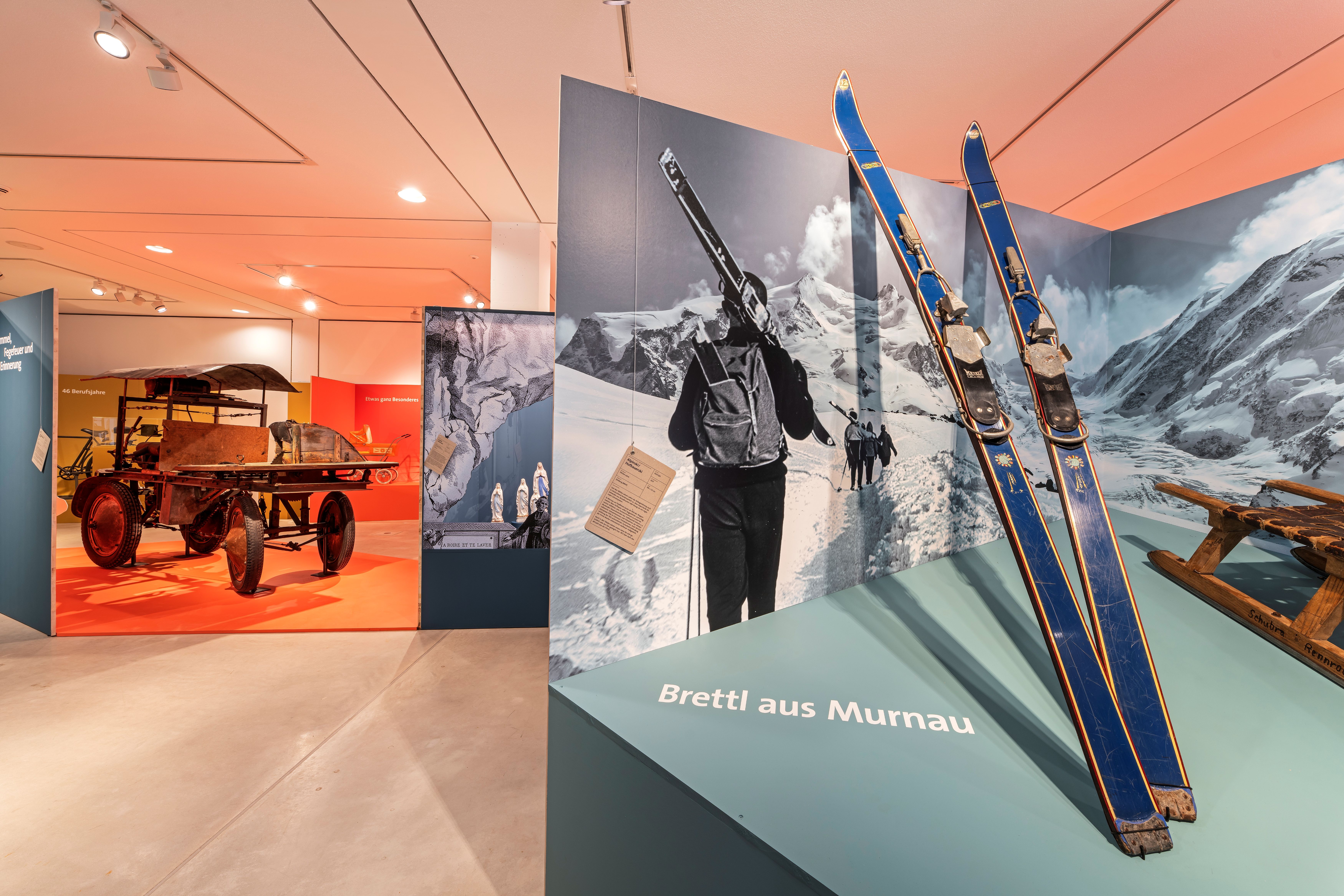 In einer Ausstellung sind verschiedene Objekte ausgestellt. Im Vordergrund werden auf einem schrägen Podest Skier präsentiert, dahinter eine mobile Kreissäge.