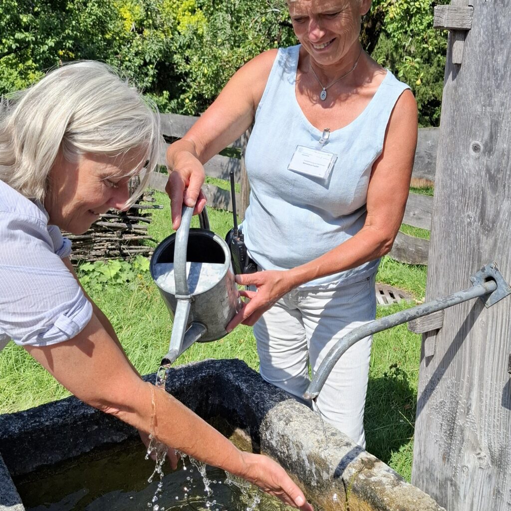 Kneippgüsse im Freilichtmuseum Glentleiten Eine Frau hält ihre Arme über einen Brunnen. Eine weitere Frau gießt mit einer Kanne Wasser über die Arme.