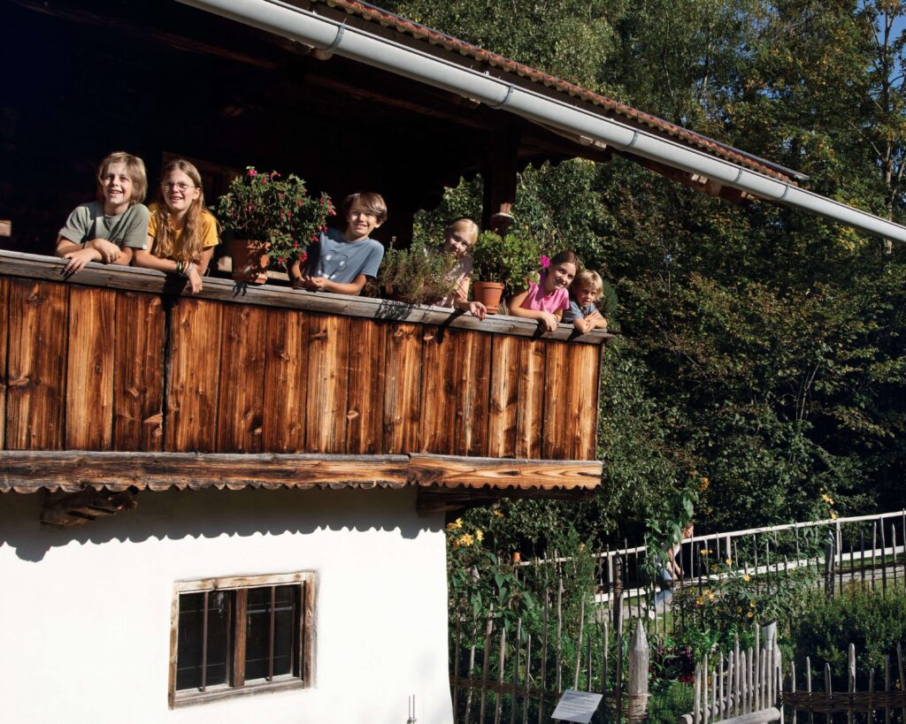 Mit Spiel und Spaß die Glentleiten erkunden Sechs Kinder lehnen sich lachend über ein Balkongeländer. Zwischen ihnen stehen Blumentöpfe.