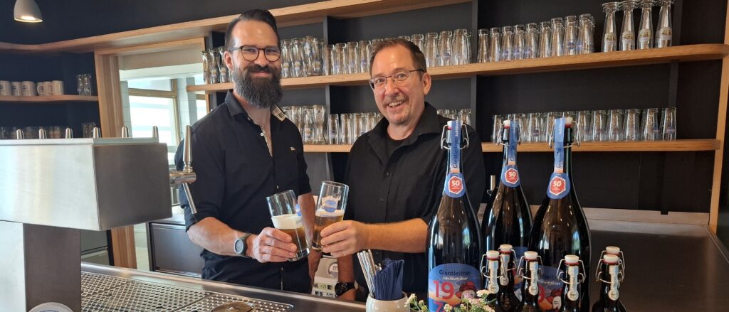 Die Wirte Sebastian Miller und Markus Pertack im Wirtshaus Glentleiten Zwei Männer stehen lächelnd hinter einem Tresen mit Zapfanlage. Sie prosten sich mit Biergläsern zu.