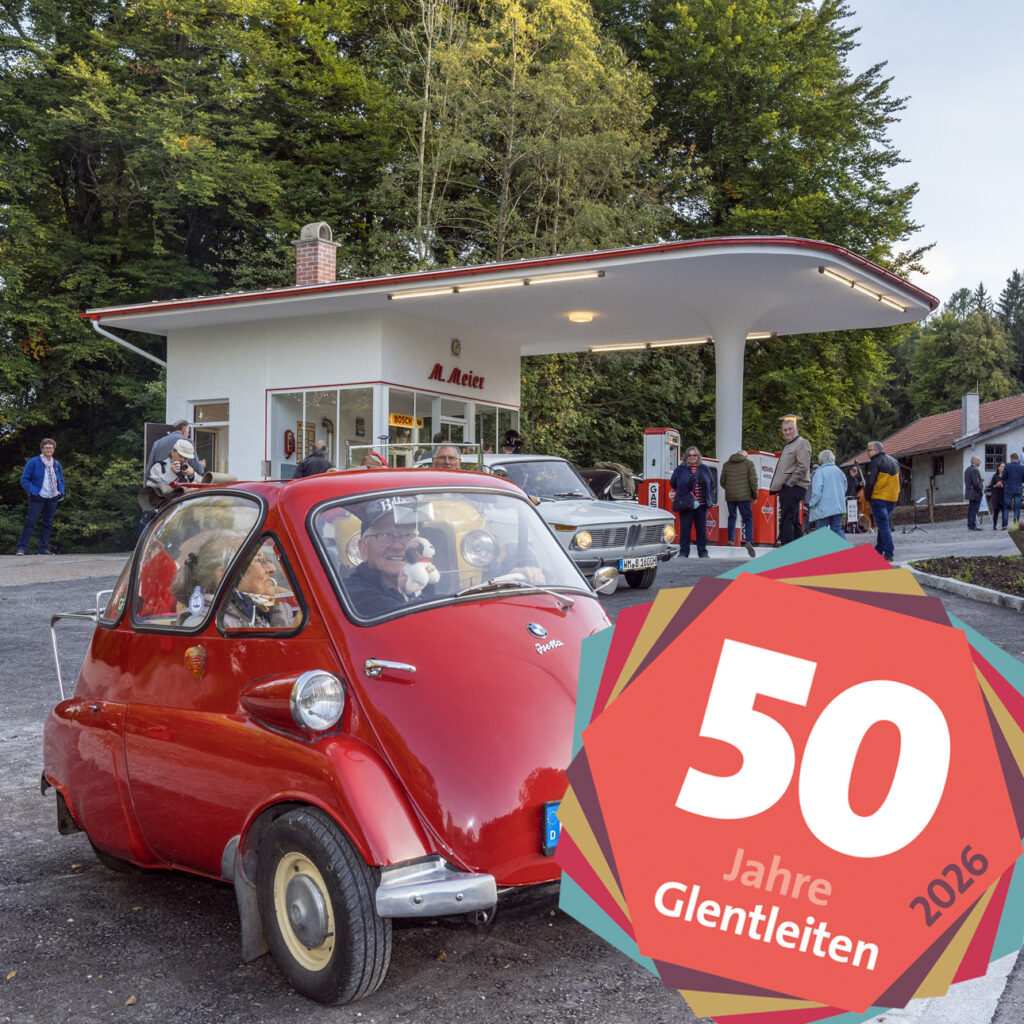 Ein rotes sehr kleines Oldtimer-Auto fahrt von einer alten Tankstelle weg.