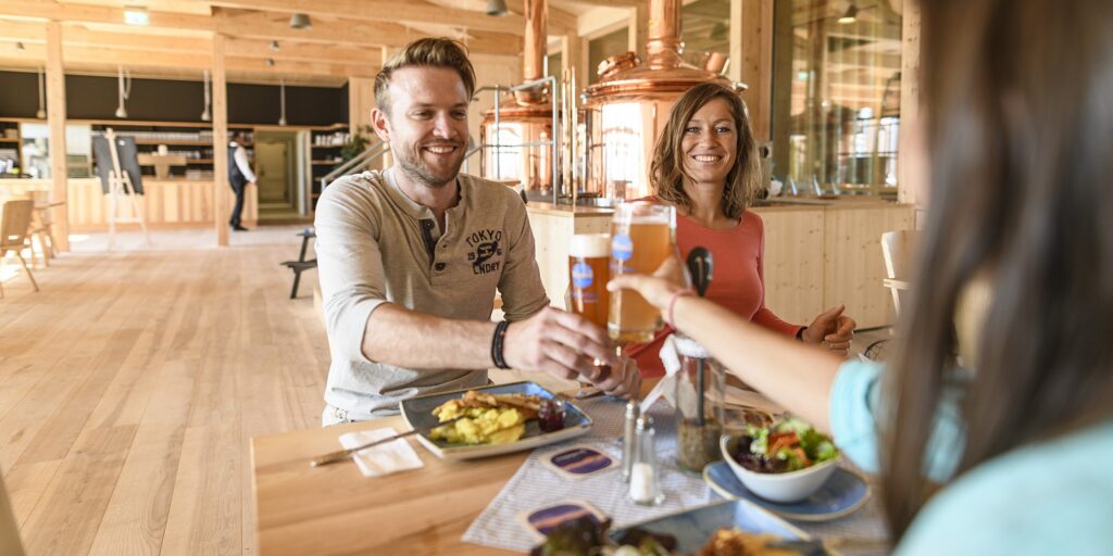 Wirtshaus Glentleiten – hier wird alpenländische Küche serviert Ein Mann und zwei Frauen sitzen an einem Tisch in einem Restaurant. Sie prosten sich mit Bier zu. Auf dem Tisch stehen Teller mit Essen.
