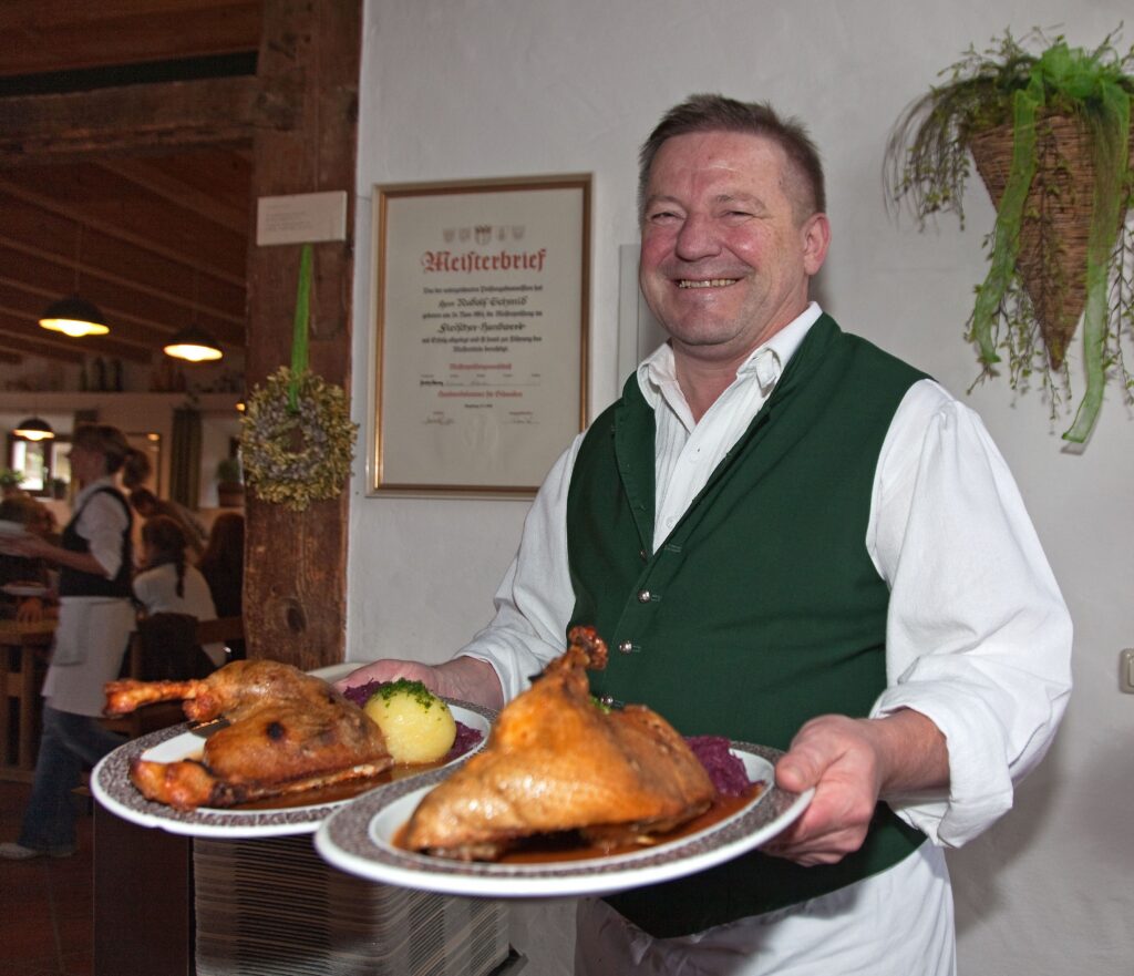 Rudi Schmid, langjähriger Wirt im Starkerer-Stadel Ein Mann in weißem Hemd und grüner Weste trägt zwei Teller mit Gänsebraten.